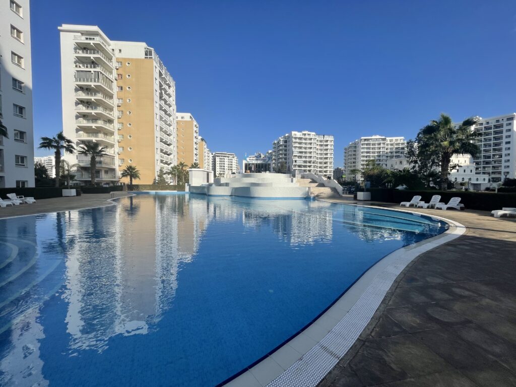 Poollandschaft mit modernen Wohngebäuden und Palmen im Hintergrund.