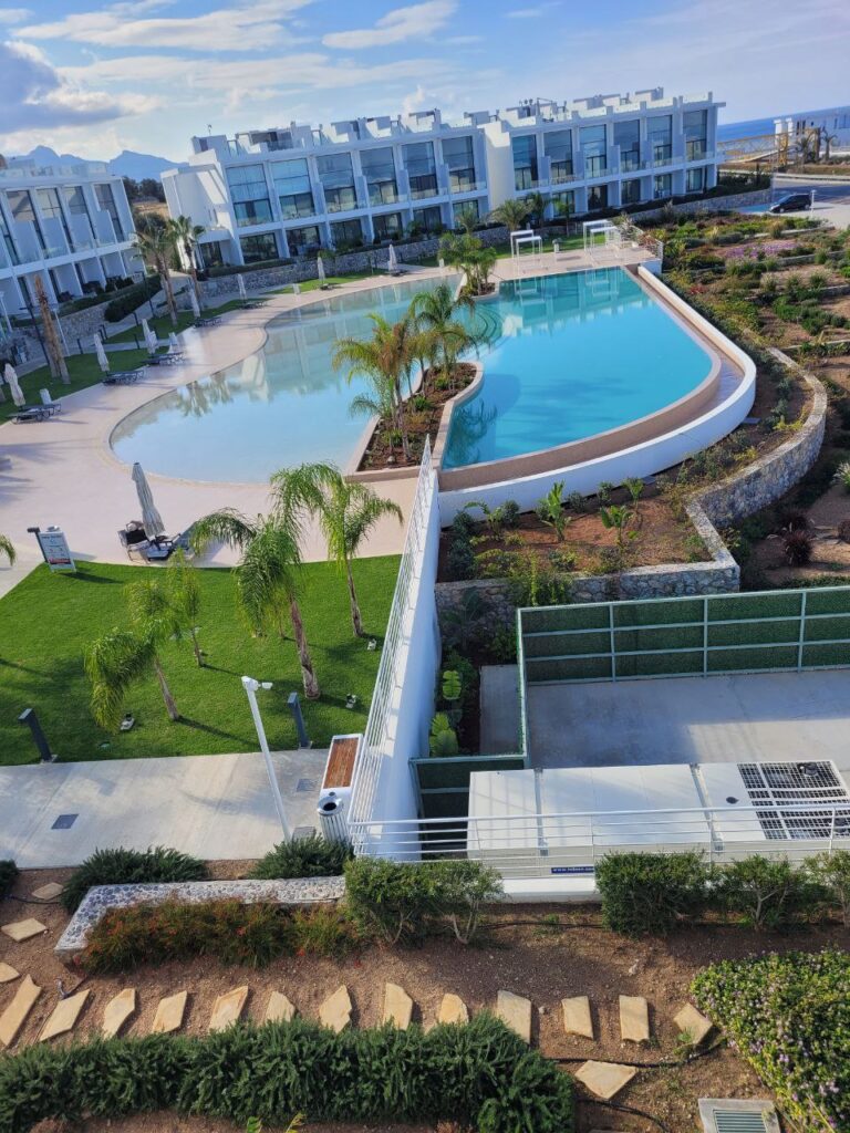 Schwimmbad mit Palmen und umliegenden Rasenflächen, von einem Balkon aus aufgenommen.