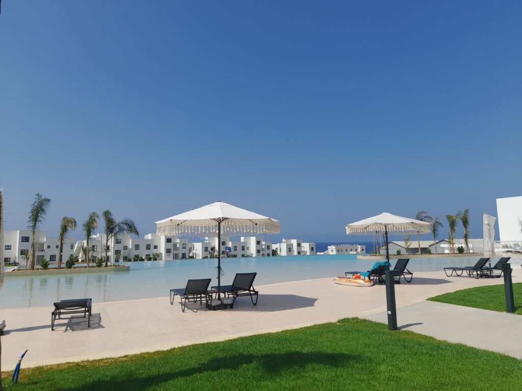 Poollandschaft mit Liegen und Sonnenschirmen, umgeben von Palmen und blauem Himmel.